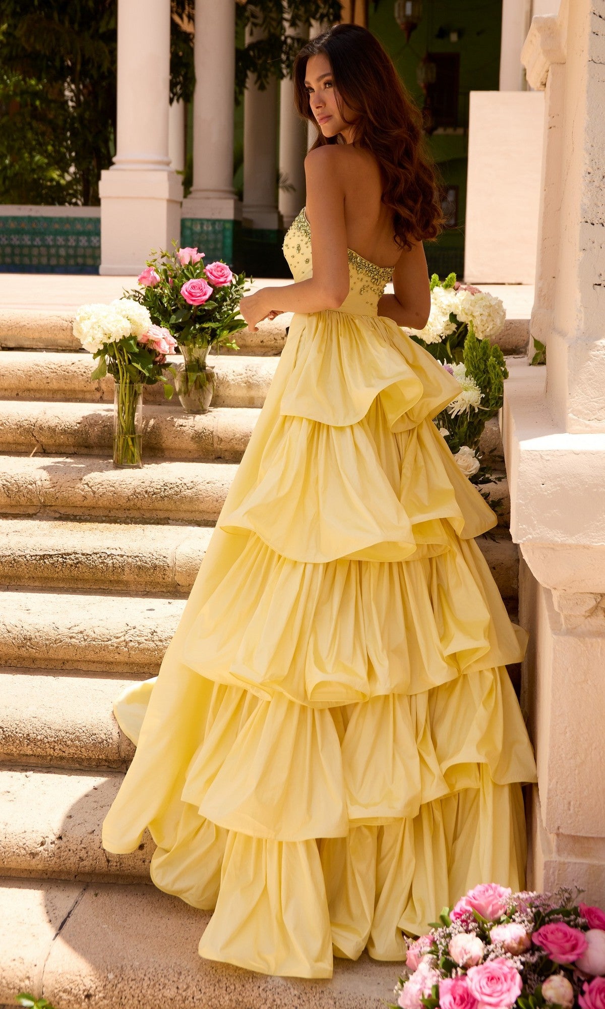 A woman in the Ava Presley 26417 long prom dress—a strapless, tiered yellow ruffle gown—stands on stone steps, holding a floral bouquet with more flower arrangements beside her.